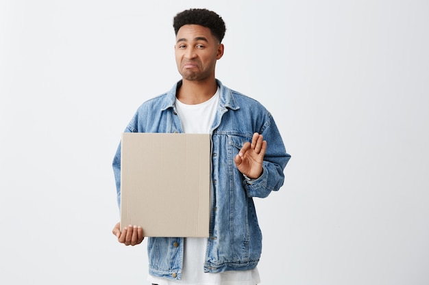 No, thanks. Close up of young funny dark-skinned american an with afro hairstyle in white t-shirt and stylish denim jacket holding clean cardboard with disgust and unsatisfied face expression