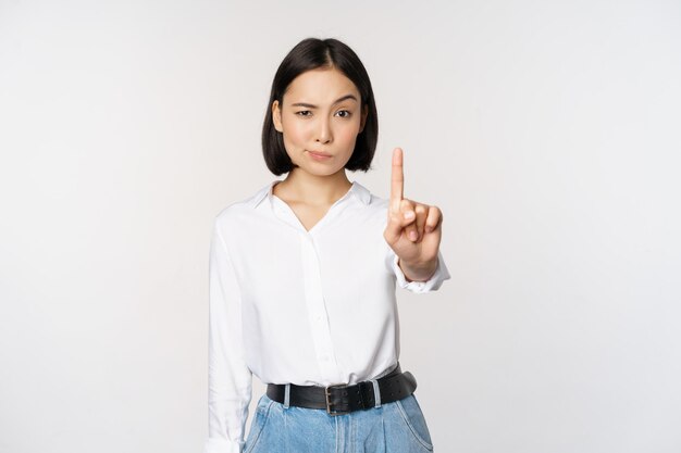 No Young serious and confident asian woman showing stop one finger gesture taboo sign standing over white background