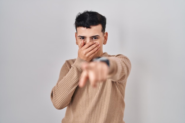 Non binary person standing over isolated background laughing at you, pointing finger to the camera with hand over mouth, shame expression