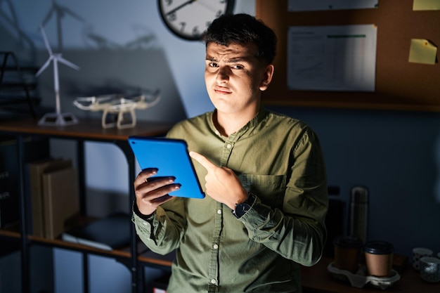 Non binary person using touchpad device at night pointing aside worried and nervous with forefinger, concerned and surprised expression