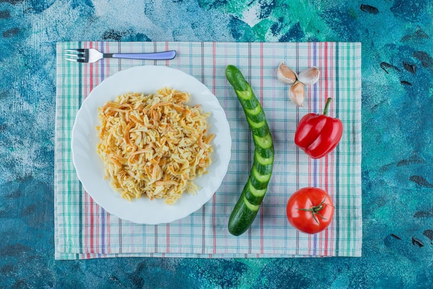 Лапша на тарелке рядом с различными овощами и вилка на кухонном полотенце на синем столе. Бесплатные Фотографии