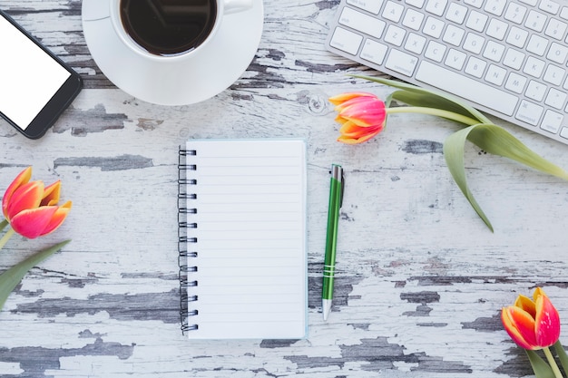 Блокнот и чашка кофе возле клавиатуры и смартфона на столе с цветами тюльпанов