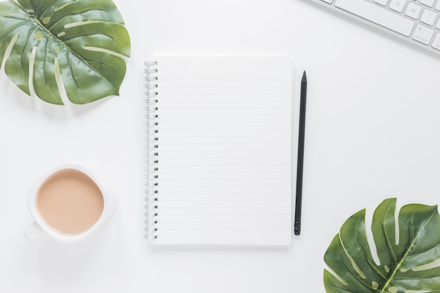 Notebook near coffee cup and keyboard on table with green leaves