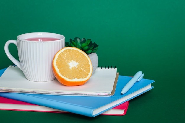 Notebooks a cup of tea and an orange on a green background copy space