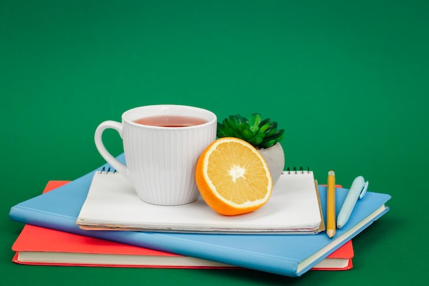 Free photo notebooks a cup of tea and an orange on a green background copy space