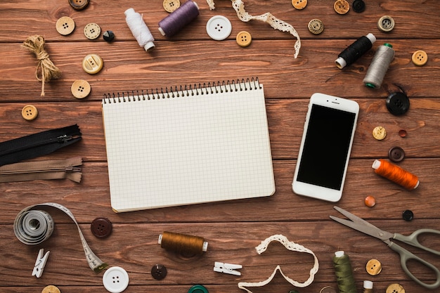 Notepad and mobile phone surrounded by sewing accessories on wooden backdrop
