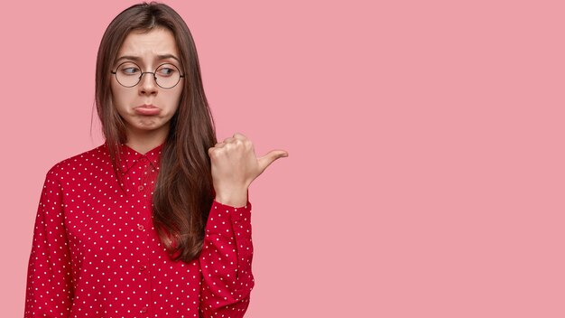 Nothing impressive there. Sad offensive woman purses lips, points right with thumb, dressed elegantly, stands over pink wall