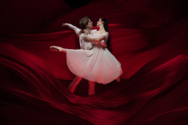 Novel. Young and graceful ballet dancers on red cloth wall in classic action. Art, motion, action, flexibility, inspiration concept. Flexible caucasian couple with billowing red waves.