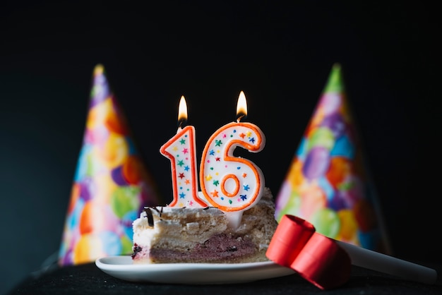 Number 16 birthday lighted candle on the slice of cake with party hat and party horn blower