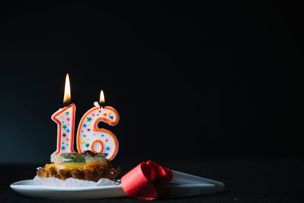 Number 16 birthday lighted candle on the slice of tart and party horn blower against black background