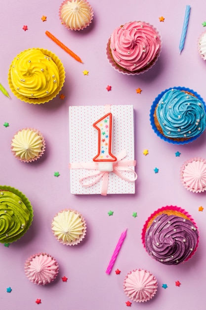 Number one candle on wrapped gift box with decorative muffins; aalaw and sprinkles on pink backdrop