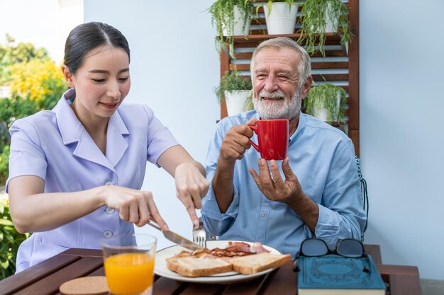 Nurse assist elderly senior man to eat breakfast and drink coffee with mug in hand at nursing home