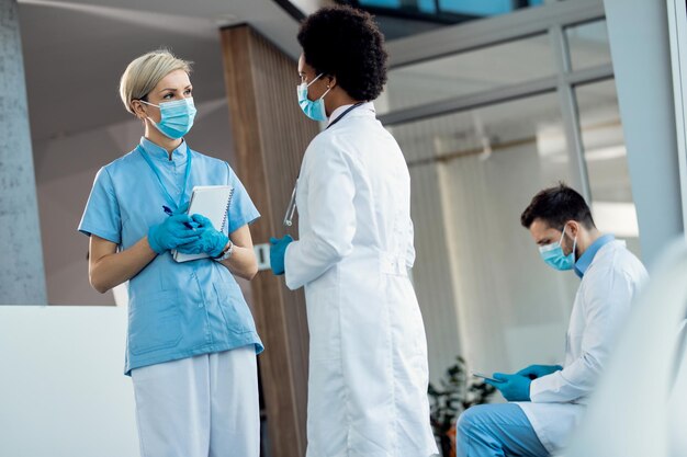 Nurse and black female doctor wearing protective face masks while talking in a hallway at the clinic