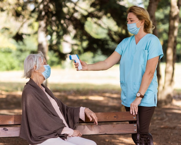 Медсестра проверяет температуру пожилой женщины на открытом воздухе на скамейке