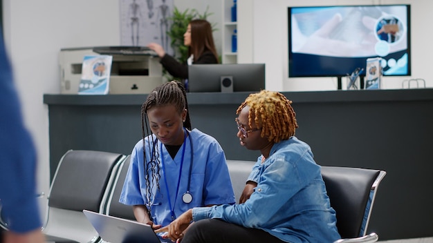 Nurse explaining diangosis on laptop to patient in waiting area at medical center. Healthcare specialist using computer at consultation exam with young adult in waiting room seats.