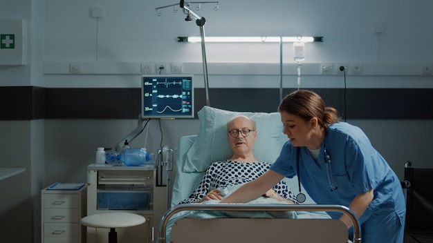 병동의 간호사가 아픈 환자를 돌보고 편안함을 위해 침대를 고정합니다. 비강 산소 튜브와 산소 측정기로 아픈 노인의 증상을 확인하는 아프리카 계 미국인 의사