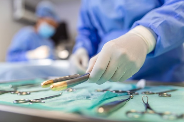 Nurse hand taking surgical instrument for group of surgeons at background operating patient in surgical theatre Steel medical instruments ready to be used Surgery and emergency concept