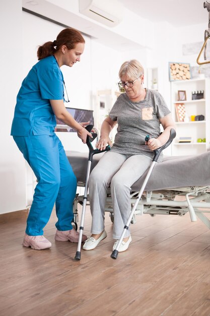 Nurse helping old woman to sit on bed in nursing home after walking with crutches.