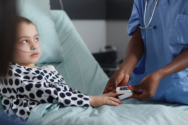 Nurse measuring oxygen levels of hospitalized sick little girl resting on patient bed. Ill kid sitting in hospital pediatric ward while medical staff monitoring health condition using oximeter.