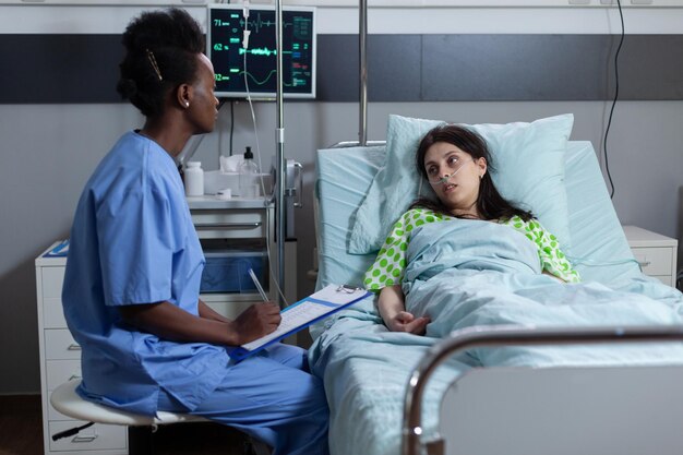 Nurse sitting at patient bedside writing on clipboard with medical chart during daily visit consulting woman with low spo2 saturation. Person on hospital bed with vitals monitored during checkup.