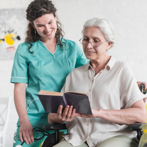 Nurse standing behind the disabled senior woman reading book