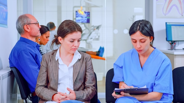 Nurse taking notes on clipboard about patient dental problems waiting for orthodontist sitting on chair in waiting room of stomatological clinic. Assistant explaining medical procedure to woman.