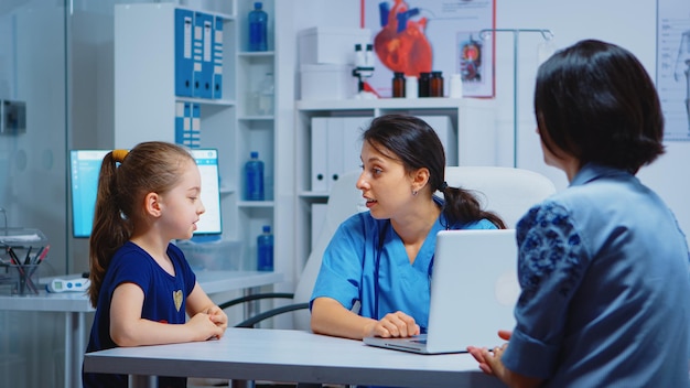 Nurse talking with child and writing girl symptoms on laptop. Physician doctor specialist in medicine providing health care services consultation diagnostic examination treatment in hospital cabinet
