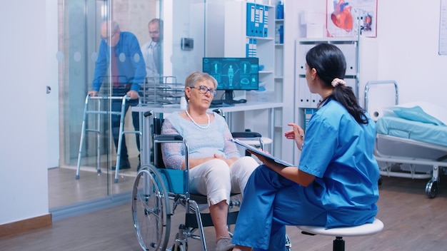 車椅子に座っている歩行障害のある年配の女性と話している看護師が、私立の近代的な回復クリニックまたは病院に行きました。障害のある老人退職患者の医療相談とアドバイス