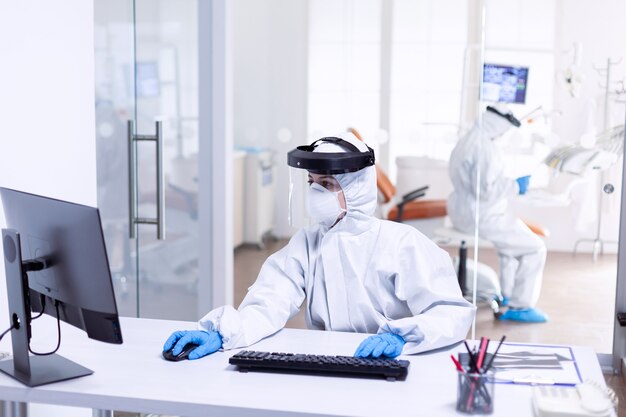 코비드 19 동안 안전 예방책으로 PPE 슈트를 입은 간호사가 컴퓨터를 사용합니다. 안전 예방책으로 치과 진료실에서 코로나바이러스 전염병에 대한 보호 장비를 착용한 의학 팀.