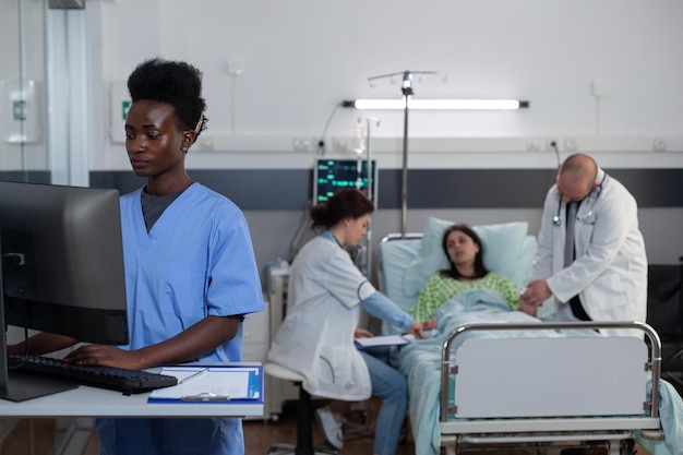 Nurse using computer to read patient medical history and lab results in hospital ward while team of doctors give clinical consult. Doctor putting pulse oximeter on patient finger checking vitals.