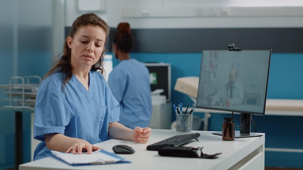 Nurse using video call for conversation with doctor on computer. Medical assistant talking to medic on online video conference for remote advice and telemedicine while sitting in cabinet