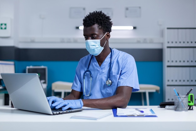 일반 개업의 캐비닛에 있는 책상에 앉아 환자 문서를 분석하기 위해 노트북을 사용하는 동안 안면 마스크를 쓴 간호사. 약의 유효기간, 성분, 복용량을 확인하는 클리닉 간호사