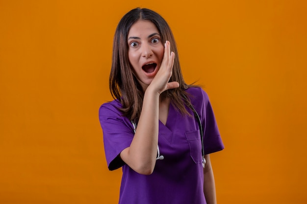 nurse wearing medical uniform and stethoscope with a hand near mouth telling a secret looking surprised standing on isolated orange