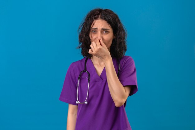 nurse wearing uniform and stethoscope closing nose with fingers feels unpleasant scent suffers from stench on isolated blue