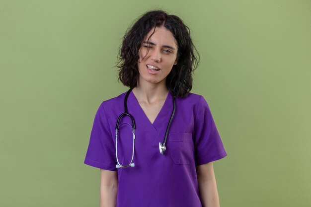 nurse wearing uniform and stethoscope confident smiling winking standing on isolated green