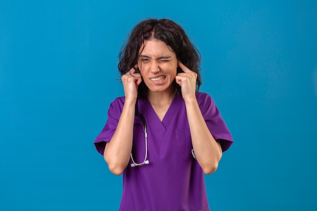 격리 된 파란색에 시끄러운 소리의 소음에 대한 성가신 표정으로 손가락으로 귀를 막고 청진기를 착용하는 간호사