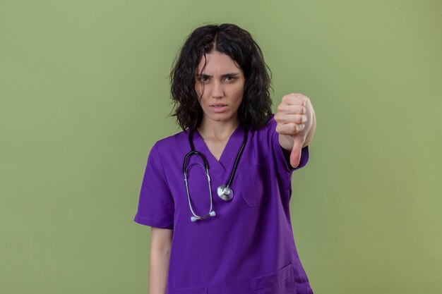 nurse wearing uniform and stethoscope displeased showing dislike thumb down standing on isolated green