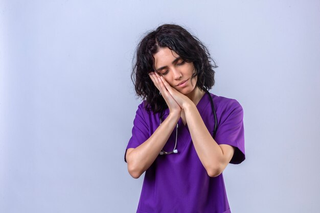 nurse wearing uniform and stethoscope looking onworked posing with hands together while smiling with closed eyes wants to sleep on isolated white