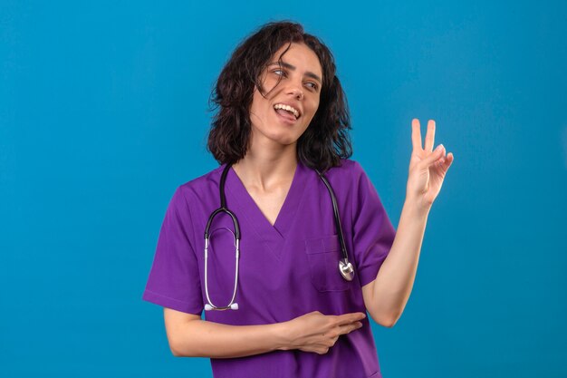 간호사 유니폼과 청진기를 입고 쾌활한 옆으로보고 웃고 고립 된 파란색에 손가락으로 두 번째 또는 승리 기호를 보여주는