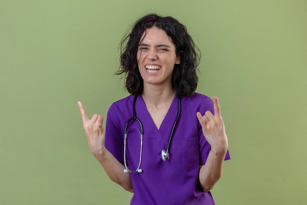 nurse wearing uniform and stethoscope smiling cheerful looking happy making rock symbols with fingers on isolated green