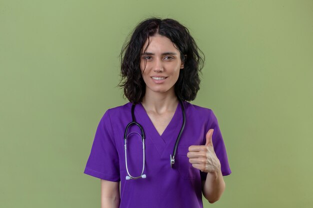 nurse wearing uniform and stethoscope with confident smile showing thumb up standing on isolated green