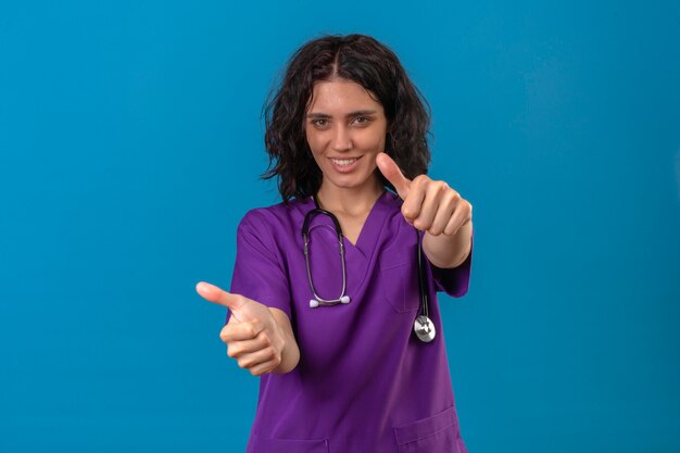 медсестра в униформе и стетоскопе с уверенной улыбкой показывает палец вверх со счастливым лицом, стоящим на изолированном синем