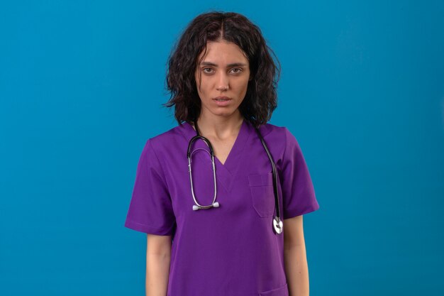 Free photo nurse wearing uniform and stethoscope with serious face without smile standing on isolated blue