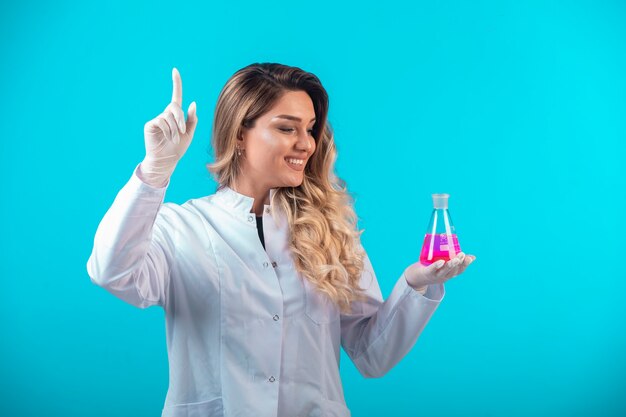 Nurse in white uniform holding a chemical flask with pink liquid and asks for attention.
