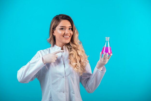 Nurse in white uniform holding a chemical flask with pink liquid and pointing at it.