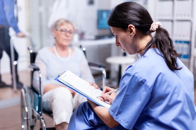 Nurse writing prescription for disabled senior woman in wheelchair after medical examination