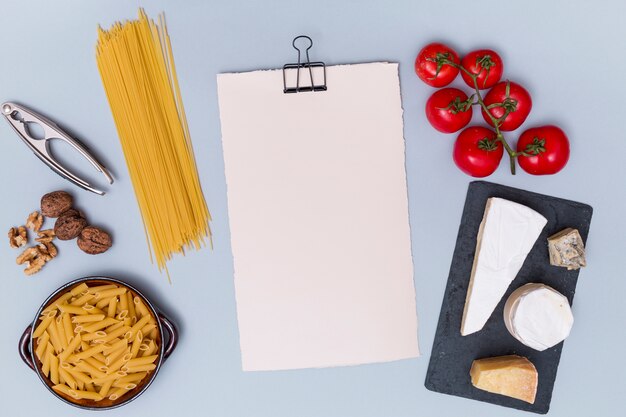 Schiaccianoci; noce; pasta cruda; vari formaggi; e pomodoro con carta bianca vuota sulla superficie grigia