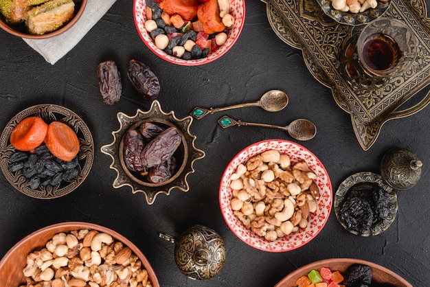 Nuts; dried fruits and dates on metallic; spoons and ceramic bowl on black background