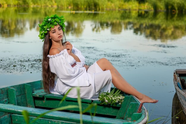 The nymph with long dark hair in a white vintage dress sitting in a boat in the middle of the river. In the hair a wreath of green leaves with white flowers. Fantasy photosession.