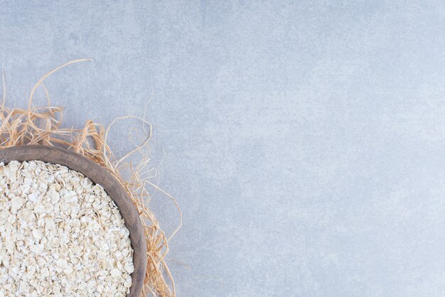 Free photo oat flakes piled in a small wooden bowl on top of a straw pile on marble background.